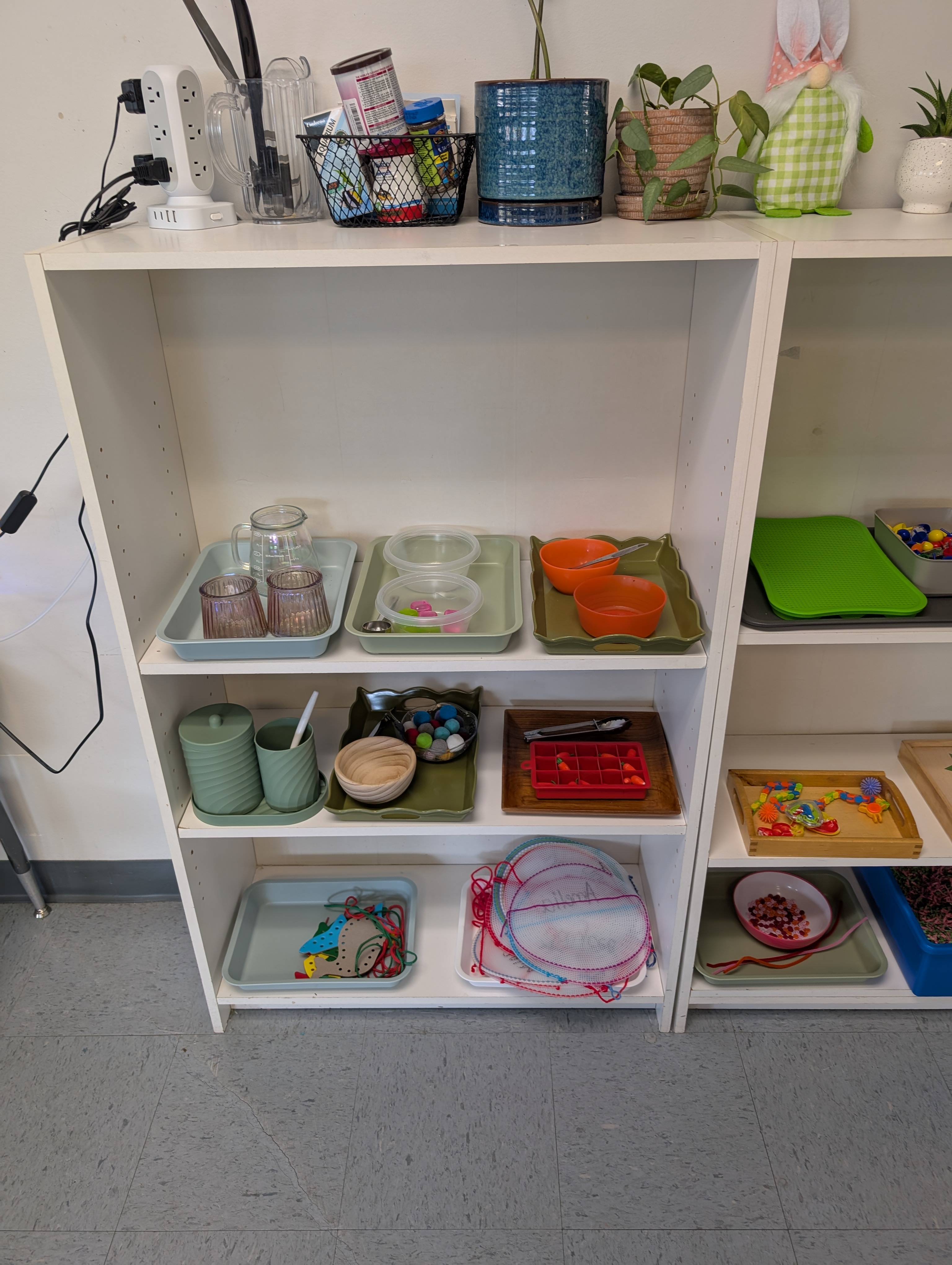 A white shelf displaying various practical life materials and tools, including cups, trays, and colorful learning objects, in a classroom setting.